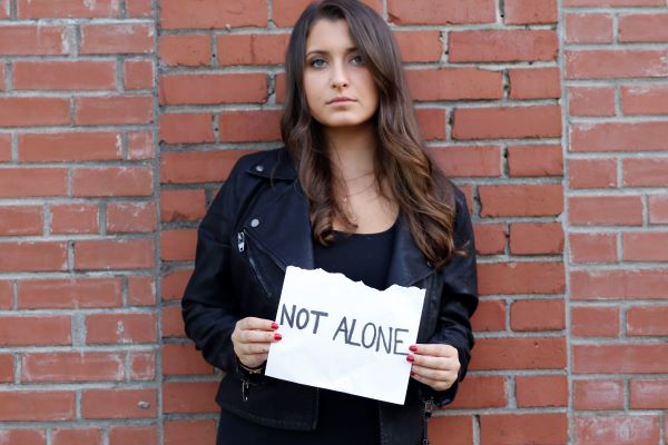 Woman holding 'NOT ALONE' sign against brick wall.